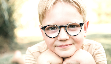 Smiling child in augie eyewear kids glasses, laying on grass cupping face with hands. 
